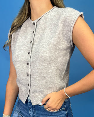 A woman wearing a light gray sleeveless button-front knit top with dark buttons, paired with blue jeans, against a solid blue background.