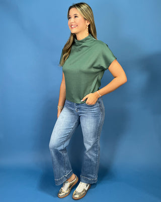 A woman modeling a sage green short-sleeve mock neck top with light-wash blue jeans and metallic sneakers, standing against a solid blue studio backdrop.