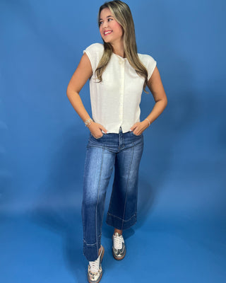 A woman modeling a white short-sleeve button-up top and blue jeans against a solid blue studio backdrop.
