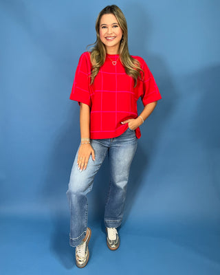 A woman modeling a bright red short-sleeve top with a white windowpane grid pattern, paired with blue jeans and white sneakers, standing against a solid blue studio backdrop.