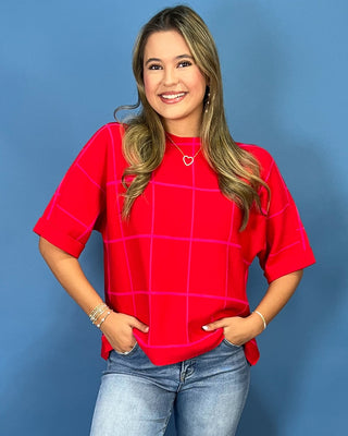 A woman wearing a bright red short-sleeve top with a pink windowpane grid pattern, paired with light-wash blue jeans