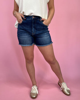Woman wearing dark-wash high-waisted denim shorts with a raw hem, styled with an ivory sleeveless top and metallic gold sneakers against a pink backdrop.