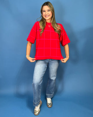 A woman modeling a bright red short-sleeve t-shirt with a thin white windowpane grid pattern, paired with blue jeans and metallic sneakers, standing against a solid blue studio backdrop.