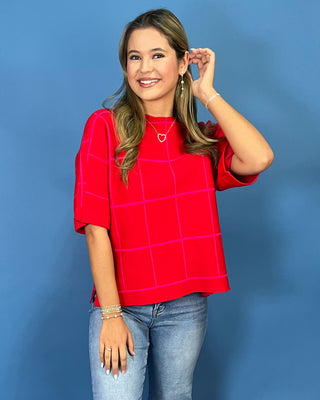 A woman modeling a bright red short-sleeve top with a pink grid pattern, paired with blue jeans, standing against a solid blue studio background.