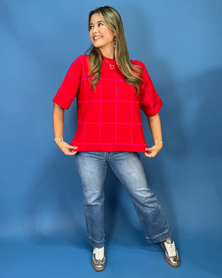 A woman modeling a bright red short-sleeve top with a white windowpane grid pattern, paired with blue jeans and metallic sneakers, standing against a solid blue studio backdrop.