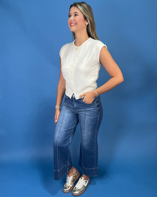A woman modeling a sleeveless cream-colored top and wide-leg blue jeans against a solid blue studio backdrop.