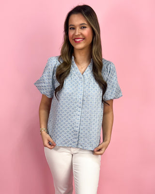 Woman wearing a blue patterned shirt and white pants against a pink background