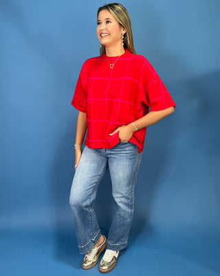 A woman modeling a bright red short-sleeve knit top with a pink windowpane plaid pattern, paired with light-wash blue jeans and metallic loafers, set against a solid blue studio backdrop.
