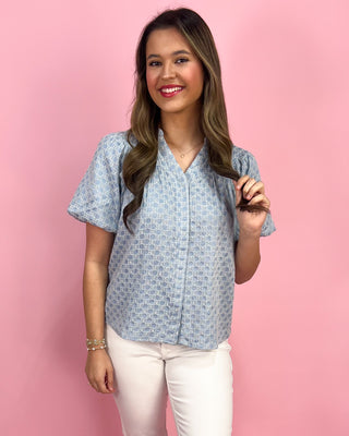 Woman wearing a blue patterned shirt and white pants against a pink background