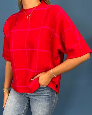 A woman wearing a bright red short-sleeve top with a magenta windowpane grid pattern, paired with light blue jeans, against a solid slate blue background.