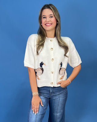 A woman modeling a cream-colored short-sleeve knit cardigan with embroidered mallards on the front, paired with blue jeans, set against a solid blue studio background.