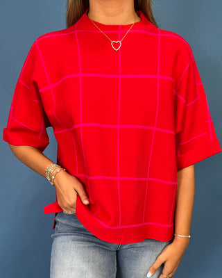 A woman wearing a bright red short-sleeve top with a pink windowpane grid pattern, paired with light blue jeans, standing against a solid blue-gray background.