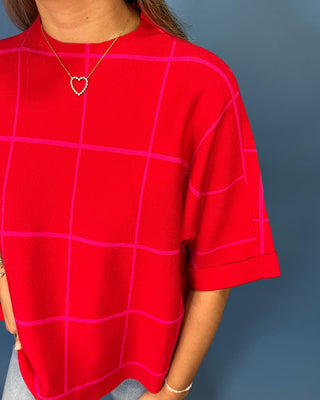 A close-up of a person wearing a bright red short-sleeve top with a pink windowpane grid pattern, paired with a gold heart necklace, set against a solid dark blue-gray background.