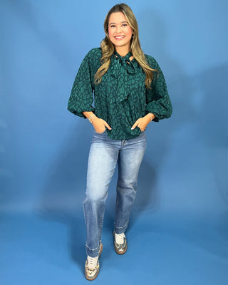 A woman modeling a dark green patterned blouse with balloon sleeves and a tie-neck detail, paired with light-wash blue jeans and metallic sneakers