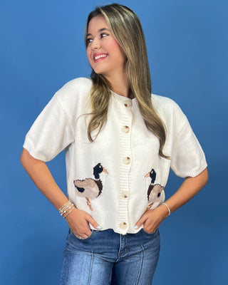 A woman wearing a cream knit cardigan with embroidered ducks on the front, paired with blue jeans, standing against a solid blue background.