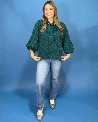 A woman modeling a dark green leopard-print blouse with three-quarter sleeves and a tie-neck, paired with light-wash blue jeans