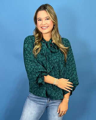 A woman modeling a dark green patterned blouse with balloon sleeves and a tie-neck detail, paired with light-wash blue jeans and metallic sneakers