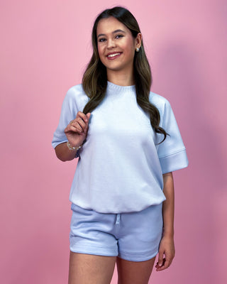 Light blue matching lounge set featuring a relaxed short sleeve top with rolled cuffs and coordinating drawstring shorts, styled with minimal jewelry against a pink backdrop.