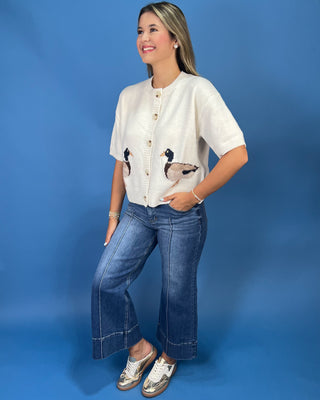 A woman modeling a short-sleeve cream-colored cardigan with embroidered ducks on the front pockets, paired with blue jeans and white sneakers, set against a solid blue studio background.