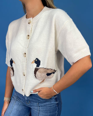 A woman wearing a cream-colored short-sleeve knit cardigan with embroidered mallards on the front, paired with blue jeans, set against a solid blue background.