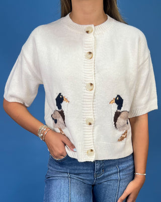 A woman wearing a cream-colored short-sleeve knit cardigan with embroidered mallards on the front and beige buttons, paired with blue jeans against a solid blue background.