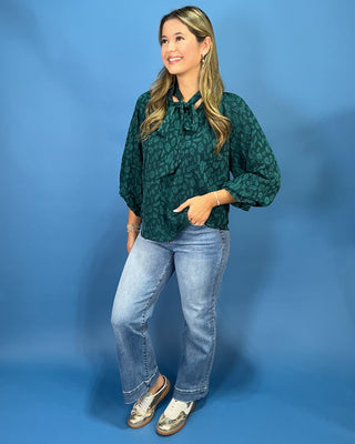 A woman modeling a dark green patterned blouse with three-quarter sleeves and a tie-neck detail, paired with light-wash jeans and metallic loafers