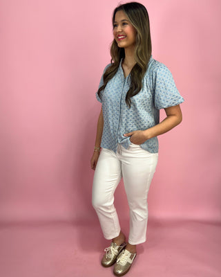 Woman wearing a light blue patterned blouse and white pants against a pink background