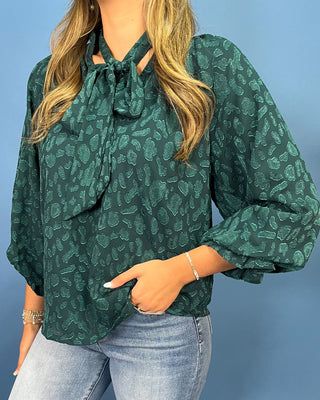 A woman wearing a dark green long-sleeve blouse with a tonal leopard print pattern, keyhole tie neckline, and puffed sleeves, paired with light blue jeans against a solid blue background.