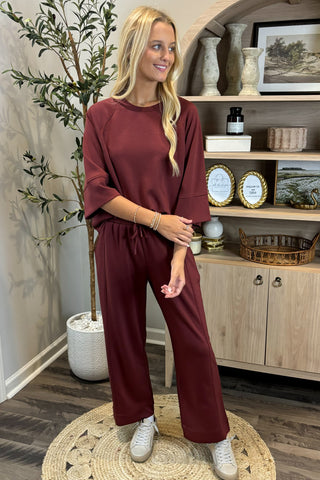 Woman in burgundy outfit standing in a room with a plant and shelves in the background