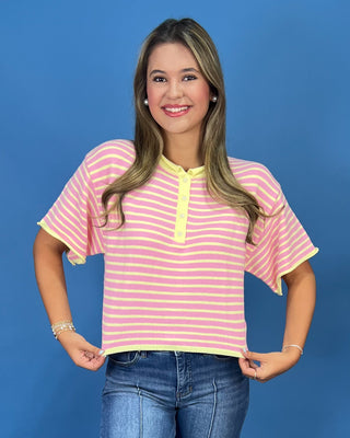 A woman modeling a pink and yellow striped short-sleeve henley top with yellow trim, paired with wide-leg blue jeans and whit