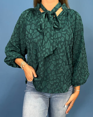 A woman wearing a dark green leopard-print blouse with a tie-neck detail and three-quarter sleeves, paired with light-wash blue jeans, set against a solid blue background.