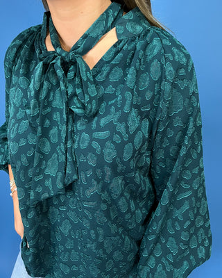 A close-up of a woman wearing a dark teal-green long-sleeve blouse with a tonal paisley pattern and a tie-neck detail, set against a solid blue background.