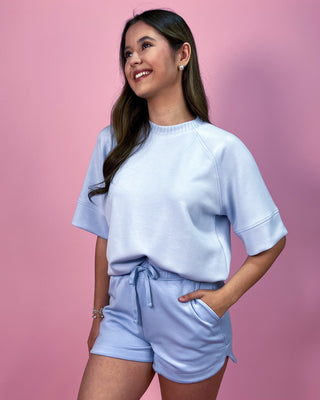 Light blue matching lounge set featuring a relaxed short sleeve top with rolled cuffs and coordinating drawstring shorts, styled with minimal jewelry against a pink backdrop.