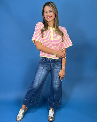 A woman modeling a pink and yellow striped short-sleeve henley top with yellow trim, paired with wide-leg blue jeans and whi
