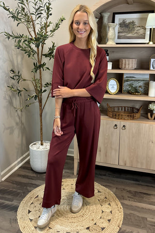 Woman in burgundy outfit standing in a room with a plant and shelves.