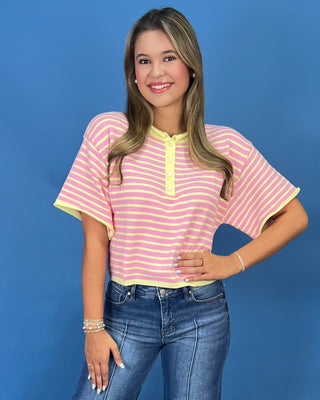 A woman modeling a pink and yellow striped short-sleeve henley top with yellow trim, paired with wide-leg blue jeans and whit