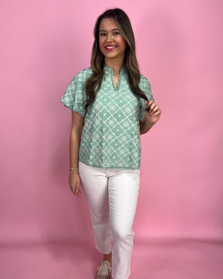 Woman wearing a green patterned top and white pants against a pink background