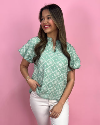 Woman wearing a green patterned blouse and white pants against a pink background