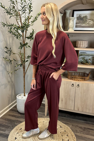 Woman in burgundy outfit standing in a room with a plant and shelves.