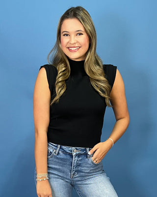 A smiling young woman with blonde hair wearing a black sleeveless mock neck top and light wash blue jeans, standing against a solid blue background.