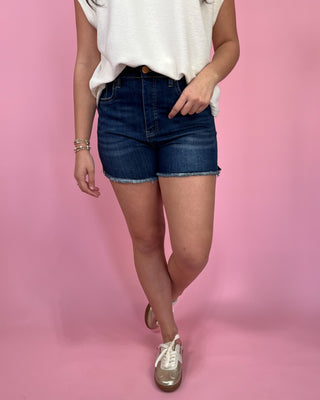 Woman wearing dark-wash high-waisted denim shorts with a raw hem, styled with an ivory sleeveless top and metallic gold sneakers against a pink backdrop.