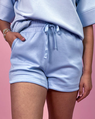 Light blue matching lounge set featuring a relaxed short sleeve top with rolled cuffs and coordinating drawstring shorts, styled with minimal jewelry against a pink backdrop.