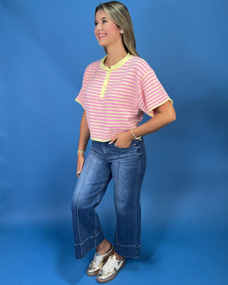 A woman modeling a pink and yellow striped short-sleeve henley top with yellow trim, paired with wide-leg blue jeans a