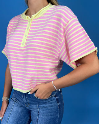 A woman modeling a pink and yellow striped short-sleeve henley top with yellow trim, paired with wide-leg blue jeans and whit