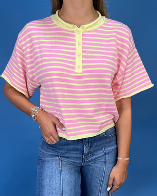 A woman wearing a pink and yellow striped short-sleeve henley top with blue jeans, standing against a solid blue background.