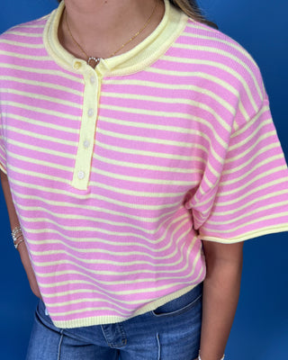 A close-up of a person wearing a pink and yellow striped short-sleeve henley top with a yellow collar and button placket,