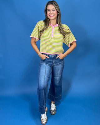A young woman modeling a yellow and green striped short-sleeve henley shirt with a pink collar trim, paired with blue jeans and white sneakers, standing against a solid blue studio backdrop.