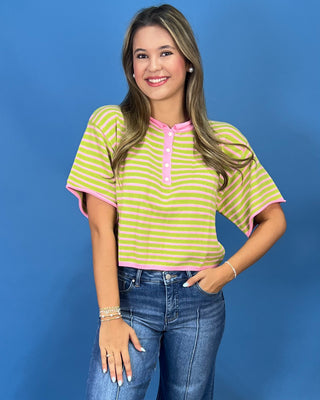 A woman modeling a short-sleeve striped henley top with lime green and white horizontal stripes and pink trim, paired with blue jeans, against a solid blue studio background.