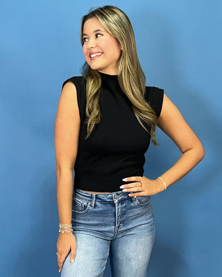 A woman with blonde-highlighted hair wearing a black sleeveless mock neck top and light-wash blue jeans, posing against a solid blue background.