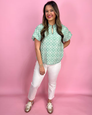 Woman wearing a green patterned top and white pants against a pink background